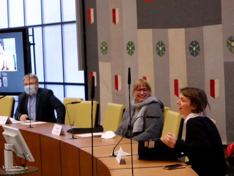 Drei Personen - zwei Frauen und ein Mann - sitzen inm Ratssaal des Neuen Rathauses hinter Pulten. Der mann lins trägt eine Maske und schaut nach vorne. Die Frau in der Mitte lacht und blickt zur Frau links, die ebenfalls lacht und nach vorne in den Ratssaal hinein blickt.